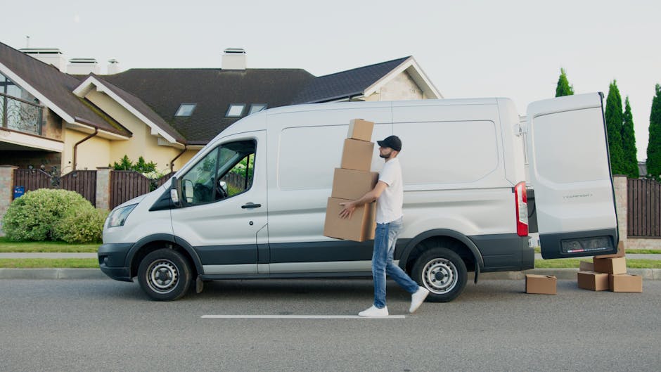 A man in casual clothing and glasses sitting on a wooden chair at the back of a white commercial van parked on a city street demonstrates the loading process involved in home relocation. The van's rea