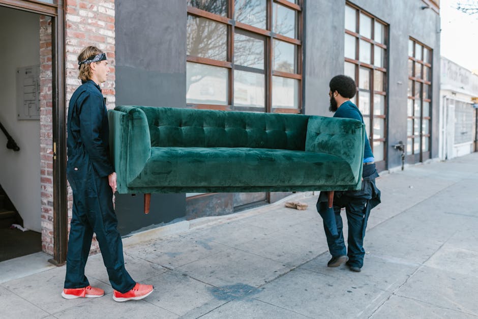 Two movers from Man with Van Camberwell are engaged in a furniture transport process outside a modern commercial building with large windows and a brick wall. The first mover, a woman wearing a dark uniform and red sneakers, is standing on the sidewalk, observing the second mover, a man with a beard dressed in a blue shirt and dark pants, who is carrying a large, green upholstered sofa with wooden legs. The sofa is being moved from inside the building to the pavement, indicating a home relocation or furniture removal activity. The environment appears well-lit with natural daylight, and there are additional structures visible down the street, emphasizing the urban setting. The scene highlights the careful handling of furniture with proper positioning and logistics involved in packing and loading on a van, supported by professional movers from Man with Van Camberwell.