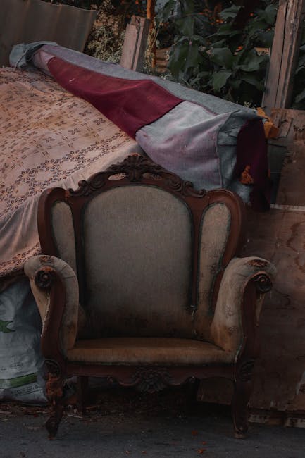 A vintage upholstered armchair with wooden carved detailing and light-colored fabric is positioned indoors on a patterned rug. Behind the chair, a large item covered with a multicolored fabric sheet is resting on a wooden surface, possibly a table or a piece of furniture. The setting appears to be a cluttered indoor space or storage area, with visible wooden beams, greenery outside through an open window or door, and various objects around. This scene shows preparations for home relocation or furniture transport, typical of professional removals services such as those provided by Man with Van Camberwell, supporting efficient packing and furniture moving procedures.