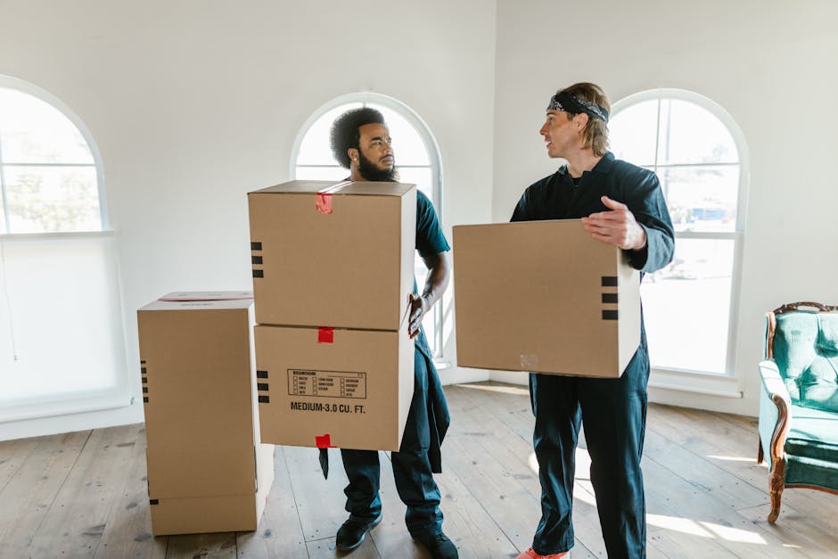 Two movers from Man with Van Camberwell are inside a bright, spacious room with white walls and large arched windows that let in natural light. One mover, with dark hair and a beard, is holding two medium-sized cardboard boxes stacked one on top of the other, with red tape securing them and printed labels visible. The other mover, wearing a headband, is holding a single cardboard box and is engaged in conversation with him. The boxes are made of sturdy cardboard, some wrapped in plastic for protection, and are positioned near an open doorway that leads to a corridor or another room. In the background, additional cardboard boxes are stacked on the wooden floor, ready for transport, indicating the process of packing and loading during a house or flat move. The scene is set in an environment prepared for a home relocation, with equipment such as trolleys, straps, or blankets possibly out of sight but implied by the context of furniture transport and moving logistics involved in the service provided by Man with Van Camberwell.