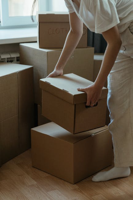 A person in casual clothing, including a beige T-shirt and light-colored trousers, is inside a room surrounded by large cardboard boxes used for packing and moving belongings. The individual is in the process of lifting and stacking a medium-sized cardboard box, which is plain with no visible markings, onto a larger box on the floor. Additional boxes, some labeled with handwritten notes such as 'CLOTH,' are situated nearby, indicating packing preparation for a home relocation. The room is well-lit with natural light coming through a window, and the overall environment appears organized for a house or flat move. The scene reflects the packing and loading stages involved in furniture transport and moving logistics, and it showcases the type of contents managed by companies like Man with Van Camberwell during a moving service.