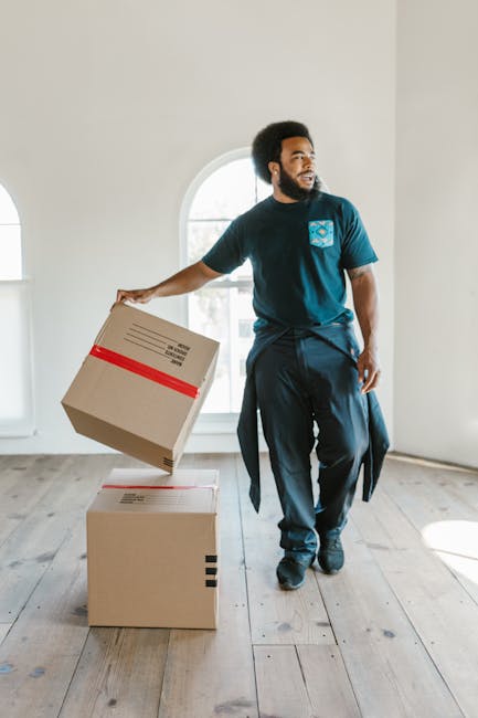 Two movers from Man with Van Camberwell are inside a bright, spacious room with white walls and large arched windows that let in natural light. One mover, with dark hair and a beard, is holding two medium-sized cardboard boxes stacked one on top of the other, with red tape securing them and printed labels visible. The other mover, wearing a headband, is holding a single cardboard box and is engaged in conversation with him. The boxes are made of sturdy cardboard, some wrapped in plastic for protection, and are positioned near an open doorway that leads to a corridor or another room. In the background, additional cardboard boxes are stacked on the wooden floor, ready for transport, indicating the process of packing and loading during a house or flat move. The scene is set in an environment prepared for a home relocation, with equipment such as trolleys, straps, or blankets possibly out of sight but implied by the context of furniture transport and moving logistics involved in the service provided by Man with Van Camberwell.