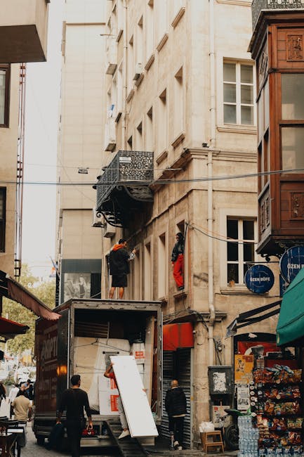 In a narrow street scene in Camberwell, a man from Man with Van Camberwell is seen unloading furniture and packed cardboard boxes from a moving van positioned at the curbside, adjacent to a multi-storey building with a small decorative balcony. The loading process involves lifting and carrying items on a pavement area lined with small shops, with some objects partially obscured by the vehicle. Nearby, a few people are walking or observing the activity, while the surrounding architecture features a mix of older stone facades and a wooden bay window structure. The scene is lit by natural daylight, highlighting the logistics of home relocation and the careful handling of household items during furniture transport for a local move within Camberwell, reflecting professional packing and moving procedures employed by [COMPANY_NAME].