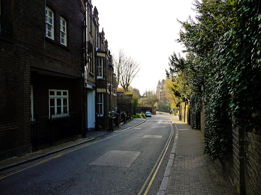 In a narrow street scene in Camberwell, a man from Man with Van Camberwell is seen unloading furniture and packed cardboard boxes from a moving van positioned at the curbside, adjacent to a multi-storey building with a small decorative balcony. The loading process involves lifting and carrying items on a pavement area lined with small shops, with some objects partially obscured by the vehicle. Nearby, a few people are walking or observing the activity, while the surrounding architecture features a mix of older stone facades and a wooden bay window structure. The scene is lit by natural daylight, highlighting the logistics of home relocation and the careful handling of household items during furniture transport for a local move within Camberwell, reflecting professional packing and moving procedures employed by [COMPANY_NAME].