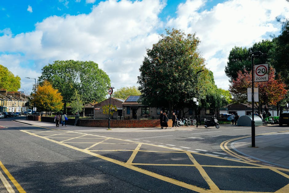 A street scene in Camberwell, Southwark, showing a residential area with a brick building surrounded by mature trees with green and orange foliage, and a sidewalk where several pedestrians are walking. In the foreground, a section of road features yellow vehicle loading and waiting restrictions, with a double yellow line and hatch markings indicating no parking zone. There is a lamppost with a 20 mph zone sign attached, and several parked bicycles and scooters are visible near the building. The sky above is partly cloudy with patches of blue. This setting depicts an urban environment suitable for house removals or furniture transport, as seen on the property doorstep and nearby loading area, with a focus on moving logistics and packing considerations for local relocation services offered by Man with Van Camberwell.