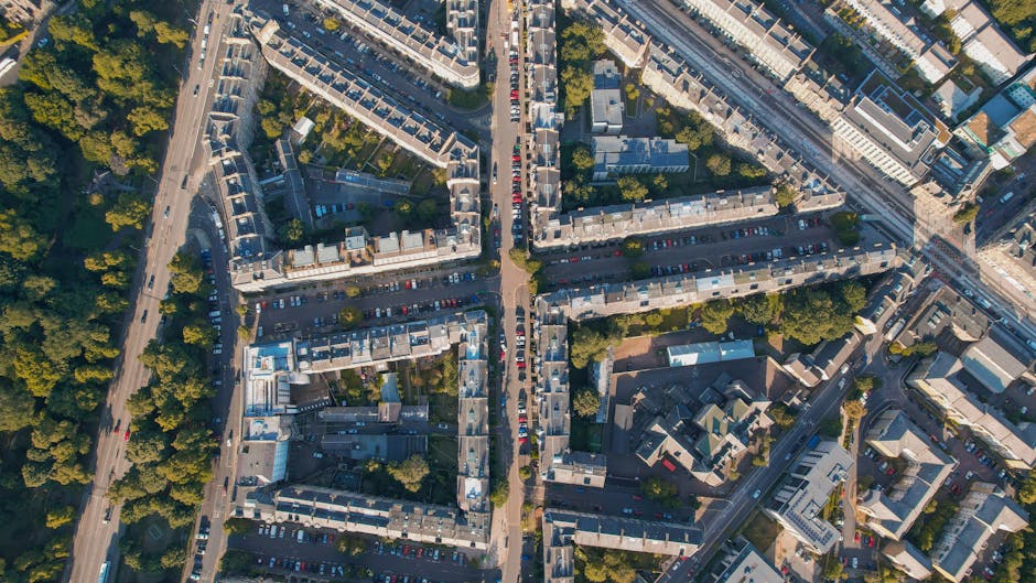 Aerial view of a densely built urban residential area showing rows of terraced houses with flat roofs, intersected by narrow streets and alleyways. In the foreground, there is a street with parked cars lining both sides, and a sidewalk with trees providing greenery. Adjacent to the houses, a larger parking lot contains numerous parked vehicles, visible beneath the branches of several trees. To the right, a section of a large modern building with a glass facade is visible, alongside other multi-story structures. The overall scene captures the character of a historic London neighbourhood, with well-maintained residential properties and a typical city arrangement of roads, parking areas, and green spaces. This image can be used to illustrate the context of property moves or home relocation in Camberwell, as managed by Man with Van Camberwell, supporting content related to house removals, packing and moving, and local moving logistics.