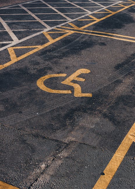 A close-up view of a designated accessible parking space featuring a yellow wheelchair symbol painted on black asphalt, with surrounding yellow diagonal lines indicating the disabled parking zone. The yellow markings are slightly worn but clearly visible, positioned adjacent to a building entrance, which is partly visible in the background. The pavement appears clean and well-maintained, with some minor cracks and patches. This scene is representative of parking areas often utilized during home relocation or furniture transport to facilitate easy access for moving services. The image is related to logistical planning for house removals, highlighting accessible parking provisions, a key aspect covered in the Camberwell New Road Removals service page provided by Man with Van Camberwell.