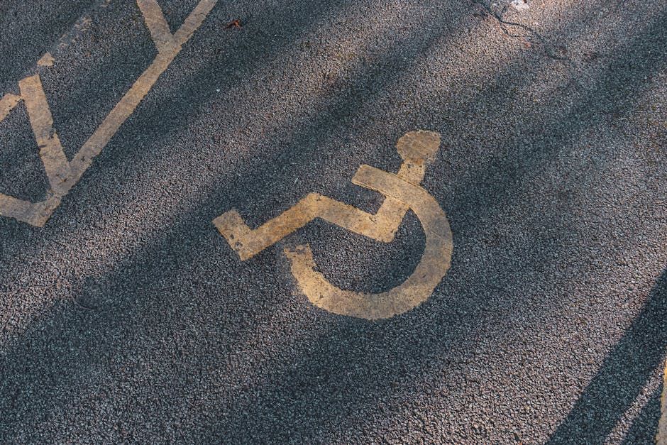 Close-up view of a disabled parking space on an asphalt surface, featuring a painted yellow wheelchair symbol with a partially visible diagonal striped area in the background. The parking lot surface shows textured asphalt with some small gravel embedded. This designated accessible parking space is likely situated adjacent to a building entrance or within a public or residential parking area, facilitating easy access for individuals with mobility needs. The image emphasizes the clear marking of the parking spot relevant to house removals or relocation services, such as those provided by Man with Van Camberwell, especially when loading or unloading furniture and boxes near a property. The surroundings suggest an outdoor parking zone under daylight conditions, with the painted lines and areas indicating regulation and designated access points for transport logistics related to home relocation projects.