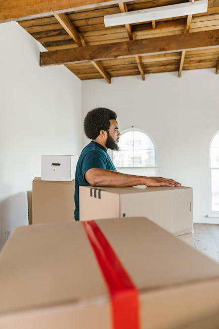 A young woman sitting cross-legged on a wooden floor surrounded by numerous cardboard moving boxes, some labeled with 'Fragile' and others plain, inside a room with a textured light blue wall. She is 
