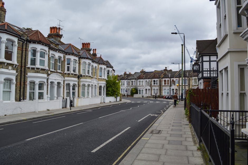 A quiet residential street with Victorian-style terraced houses featuring bay windows, decorative brickwork, and chimneys, under an overcast sky. The pavement is lined with black metal railings and concrete slabs, with a few pedestrians walking along. The road has marked parking bays and a single double yellow line, with some parked cars visible in the distance. Streetlights and a crane can be seen further along the street. This scene illustrates a typical UK neighbourhood suitable for house removals, with no visible vehicles actively loading or unloading, though it reflects the environment where transportation and packing for a home relocation by Man with Van Camberwell could take place.
