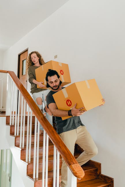 Two movers from Man with Van Camberwell are carrying cardboard boxes during a house relocation inside a residential property. The male mover, dressed in a dark grey T-shirt and beige trousers, is descending a staircase with a large cardboard box securely held in both hands. The female mover, wearing a brown top and ripped jeans, is positioned behind him on the staircase, also carrying a cardboard box. The staircase has wooden steps and a white balustrade with a polished wooden handrail. The interior features a white wall and a wooden door visible on the landing of the stairs. The environment suggests an organized moving process with careful handling of packing materials. The scene captures a professional loading process within a home, reflecting furniture transport and packing activities associated with house removals.