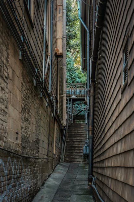 A narrow outdoor alleyway between two tall building facades, one with weathered brickwork and graffiti, the other with wooden cladding. Metal pipes and cables run along the walls, and a metal staircase with steps and a railing ascends at the end of the alley, leading to a small platform with a rounded railing. The ground is paved with concrete slabs, and the scene is lit with natural daylight, highlighting the confined space often encountered during house removals or furniture transport in tight urban areas. This setting illustrates the logistical challenge of navigating narrow staircases and alleyways when moving large items, as handled by professional removals services like Man with Van Camberwell.