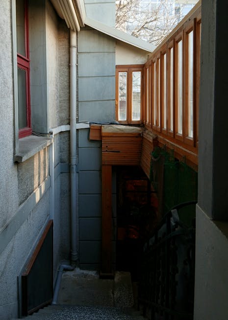 Two movers from Man with Van Camberwell are carrying cardboard boxes during a house relocation inside a residential property. The male mover, dressed in a dark grey T-shirt and beige trousers, is descending a staircase with a large cardboard box securely held in both hands. The female mover, wearing a brown top and ripped jeans, is positioned behind him on the staircase, also carrying a cardboard box. The staircase has wooden steps and a white balustrade with a polished wooden handrail. The interior features a white wall and a wooden door visible on the landing of the stairs. The environment suggests an organized moving process with careful handling of packing materials. The scene captures a professional loading process within a home, reflecting furniture transport and packing activities associated with house removals.