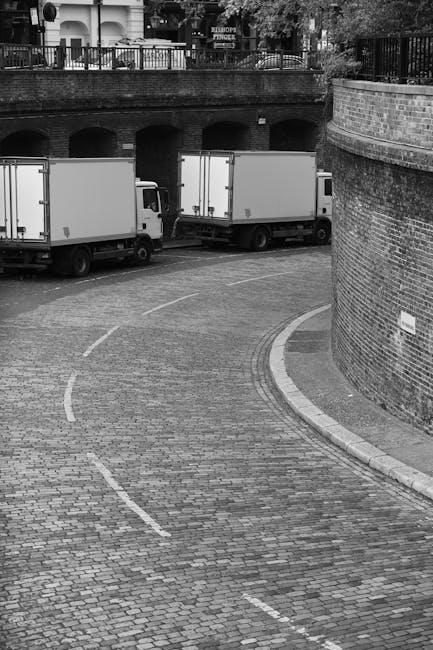 A black-and-white photograph showing two white box trucks parked on a curved, cobblestone street in an urban residential area. The trucks are positioned adjacent to a brick wall and beneath a raised area with a metal railing, which appears to be part of a balcony or walkway. The street is empty aside from the trucks, with no visible pedestrians or other vehicles. The trucks are situated near an entrance to an underground parking or service area, as indicated by the arched openings in the brick wall. The scene depicts a typical setting for home relocation or moving services, with the trucks ready for furniture transport and packing materials, including cardboard boxes and plastic-wrapped items, possibly loaded onto the vehicles nearby. The lighting is natural, with shadows cast on the cobblestones, emphasizing the textures of the street surface and the brickwork. This image illustrates the logistics process involved in professional removals, such as those offered by Man with Van Camberwell, within a residential environment with local restrictions on loading and parking.
