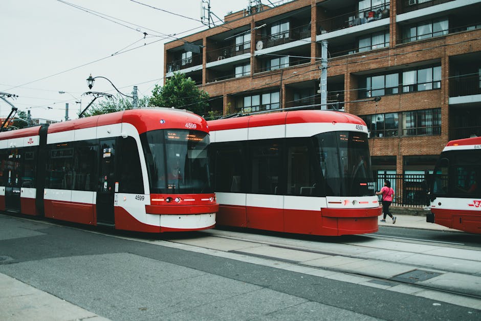 Two modern red and white trams parked on tracks along a city street with overhead electrical wires, situated in front of a multi-storey brick residential building with balconies and large windows. A person in a pink jacket is walking near the trams, and another tram is partially visible on the right. The scene is outdoors with natural daylight, showcasing urban transport infrastructure relevant to house removals and furniture transport logistics, as seen in services provided by Man with Van Camberwell.